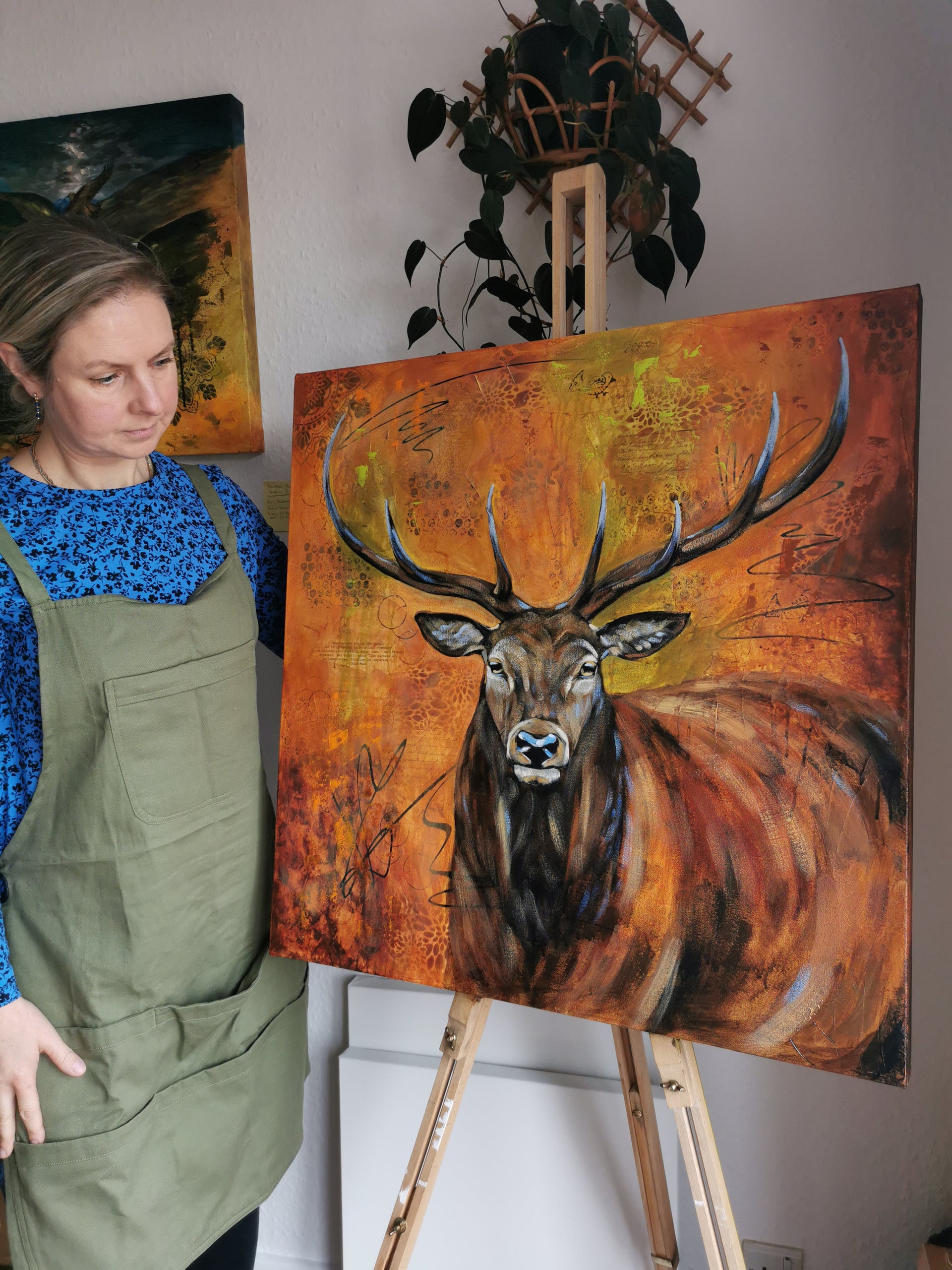 Artist in green apron standing next to a painting of a deer on an easel.