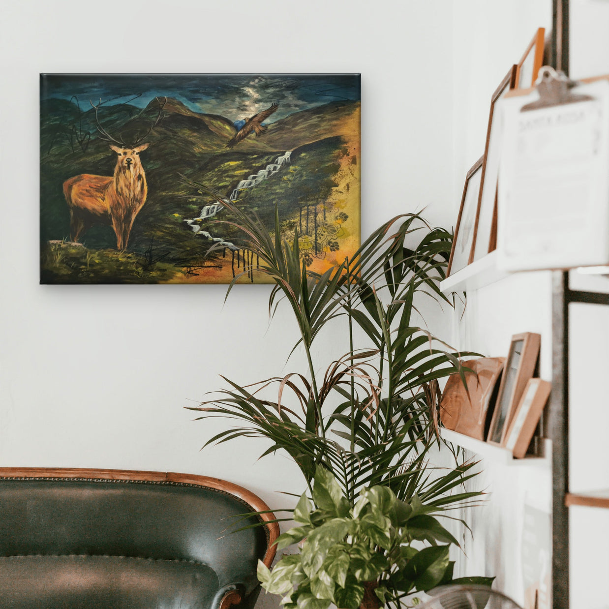 A room with a sofa, a bookshelf, and a plants featuring a painting of a stag and dramatic Scottish landscape